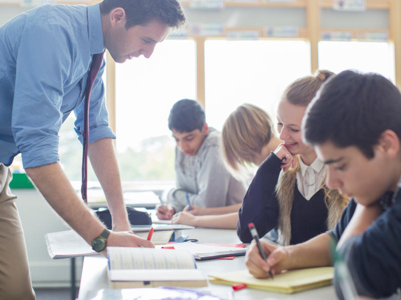 A diverse group of students working collaboratively at a table while a male teacher in business casual attire leans over to provide guidance. The students, representing different genders and ethnicities, are engaged in their work with notebooks and writing materials.