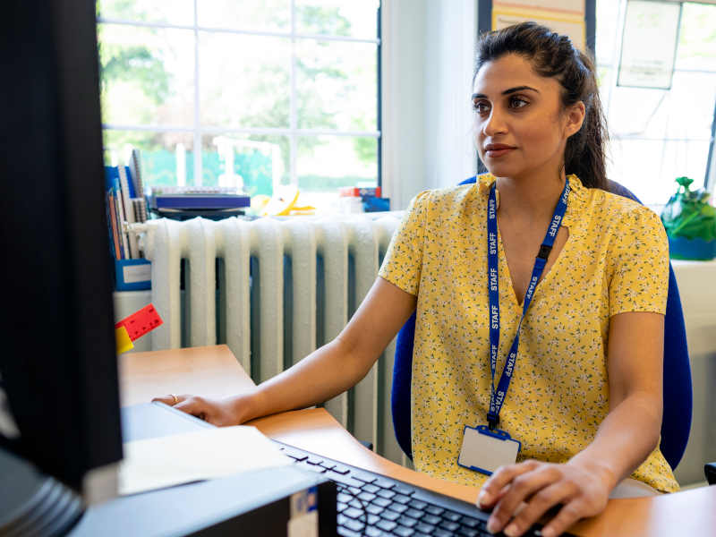 A female teacher of colour wearing a yellow floral blouse and ID badge on a lanyard works at a desktop computer, reviewing educational resources such as presentation slides or printable exercises at her desk.